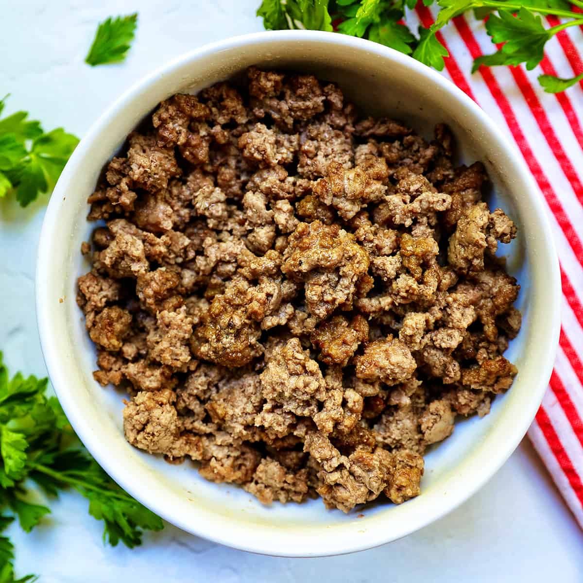 cooked ground beef in a white bowl with parsley sprigs around it and a red and white striped towel.