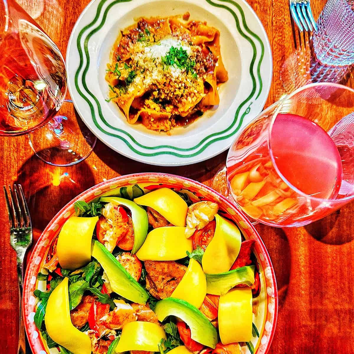 large bowl of lobster mango salad, bowl of pappardelle ragu pasta, and two glasses of wine at Bad Roman restaurant in New York City.
