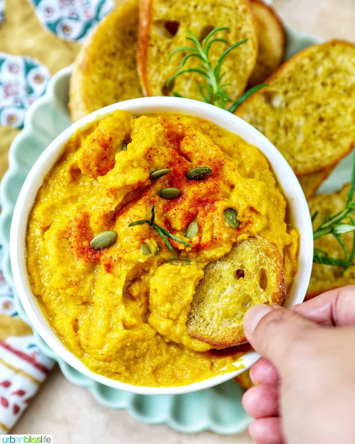hand dipping crostini into bowl of butternut squash dip with baguette slices, fresh herbs, and a colorful napkin.