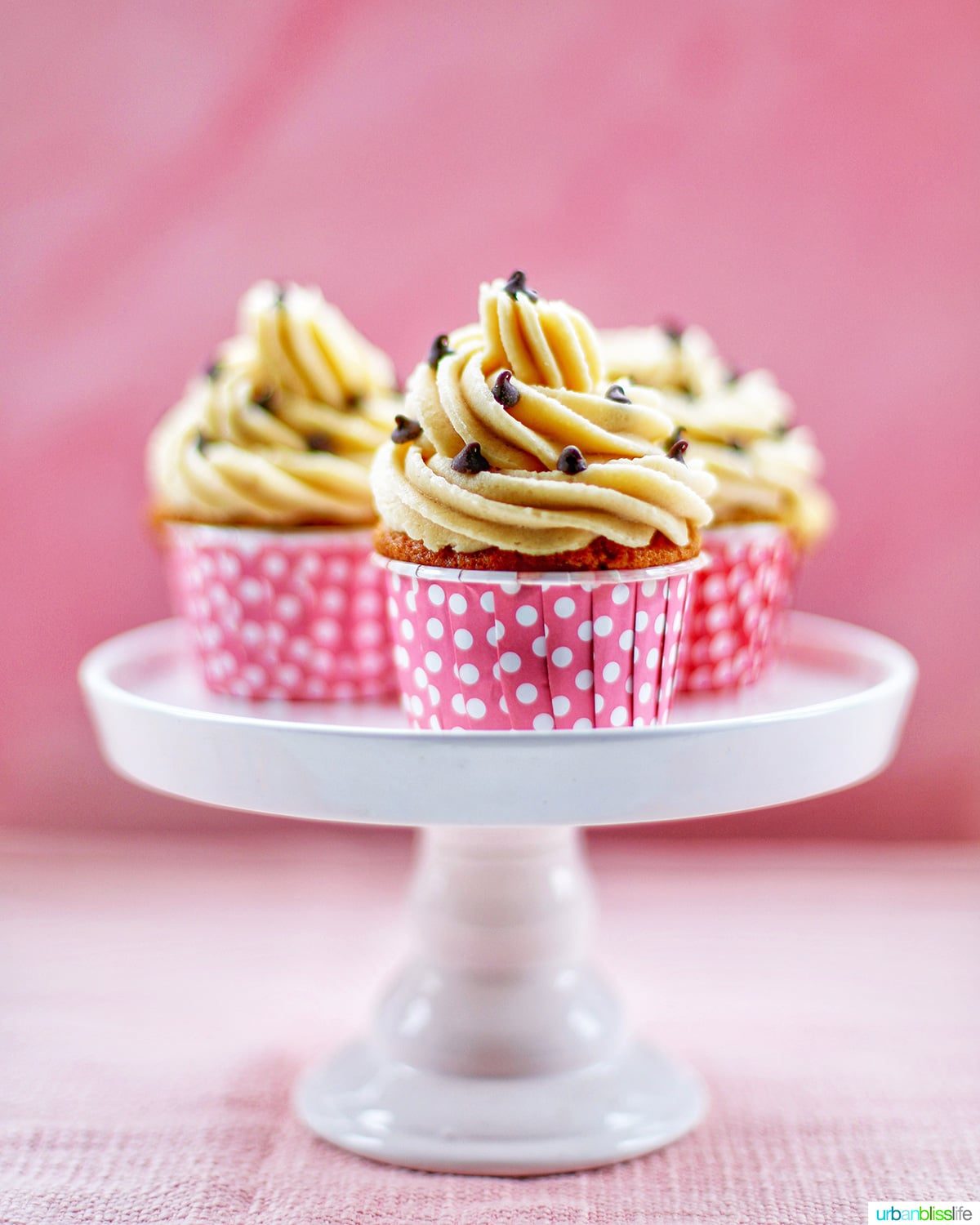 Three Chocolate Chip Cupcakes with Cookie Dough Frosting on a white cake pedestal with pink background