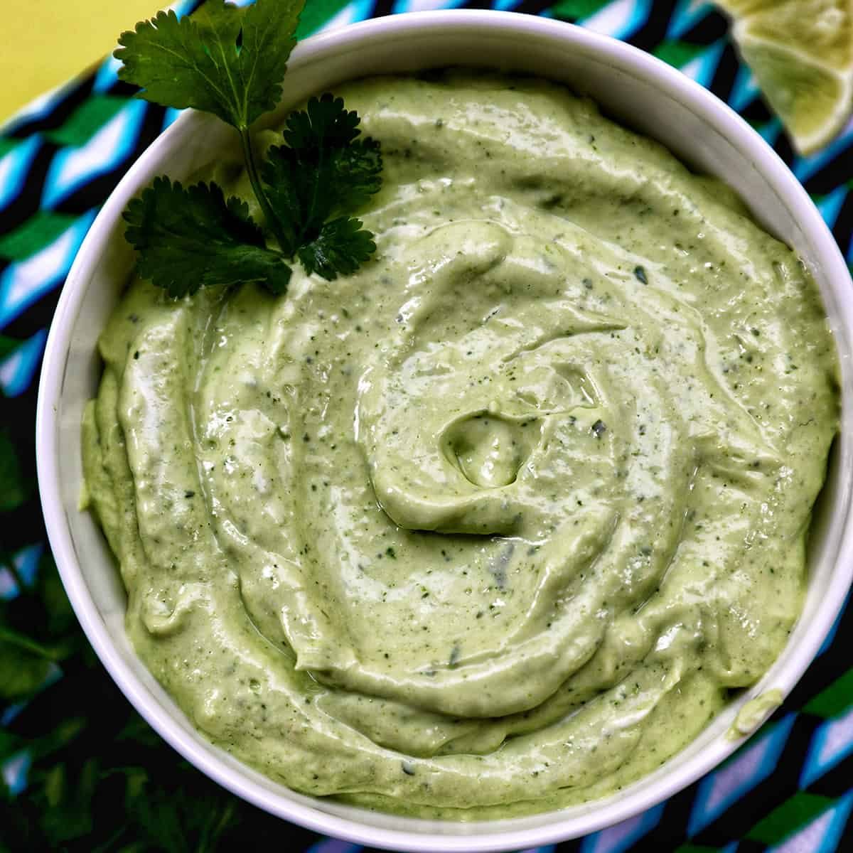 bowl of avocado cilantro lime crema on a colorful modern napkin with garnish of cilantro leaves.