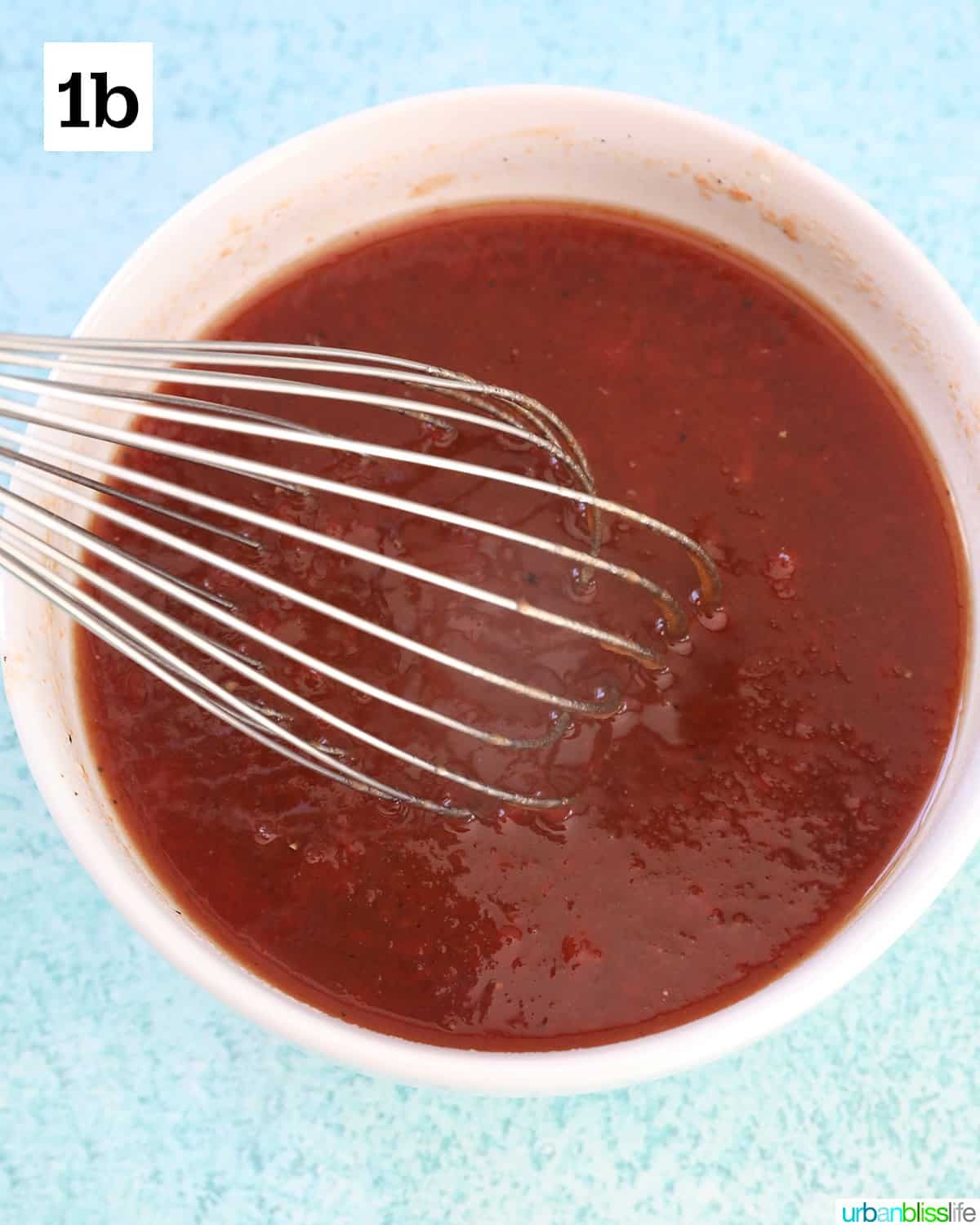 white bowl with sauce for honey bourbon meatballs and a whisk in the bowl, on top of a blue background.