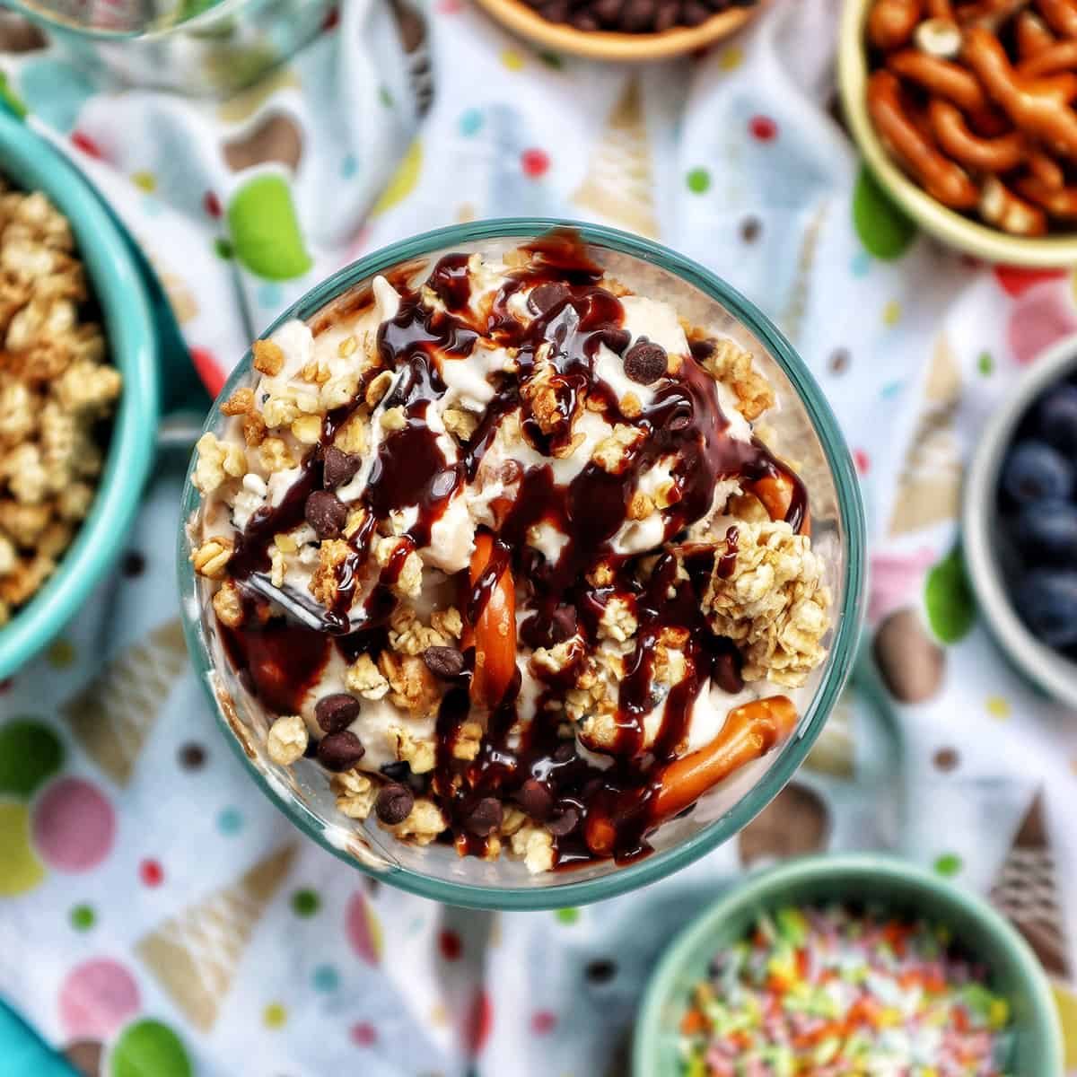 ice cream parfait topped with granola, chocolate syrup, mini chocolate chips on a colorful kitchen towel surrounded by sides of more