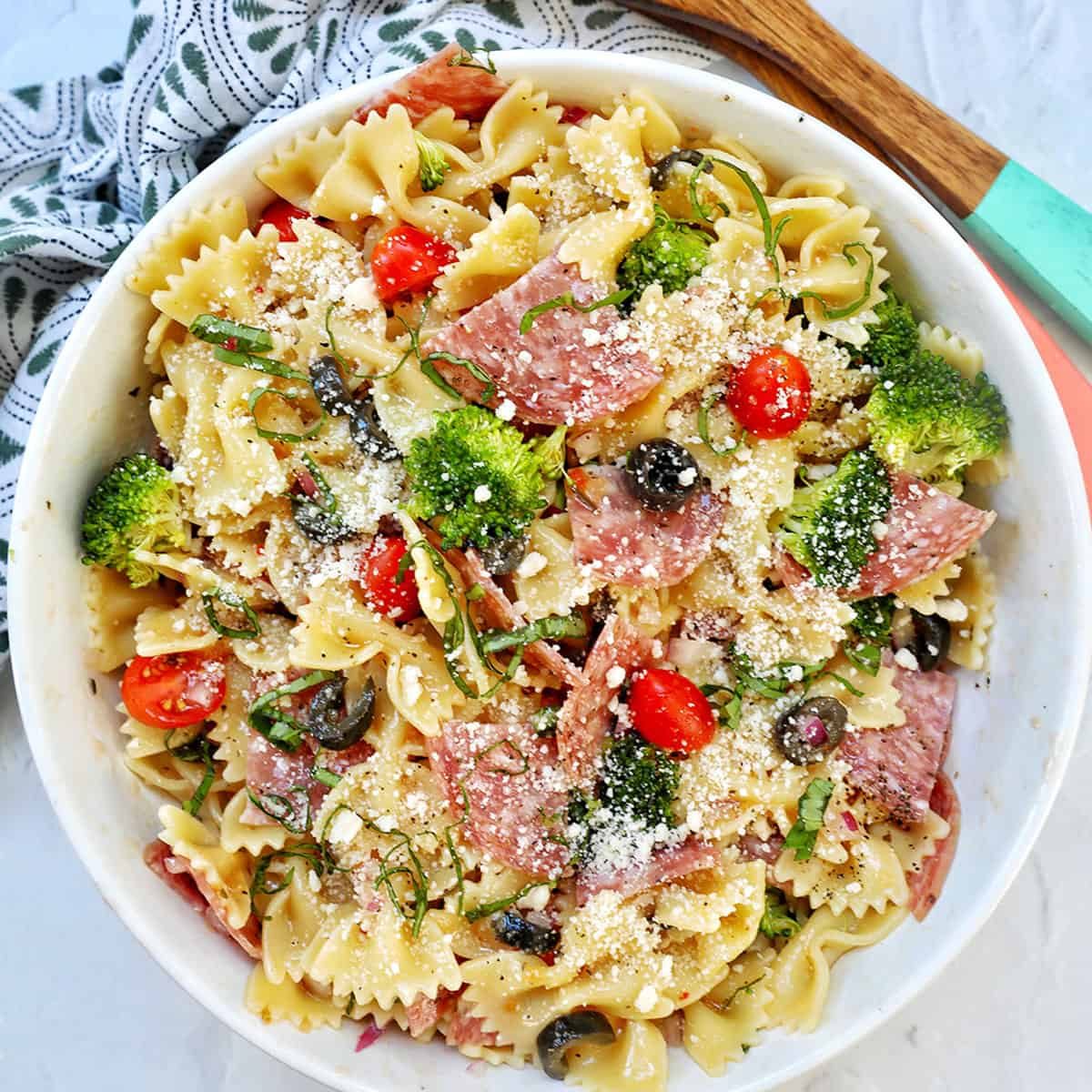 Italian bowtie pasta salad with salami, olives, tomatoes, broccoli, and parmesan cheese in a white bowl with title text that reads "Italian