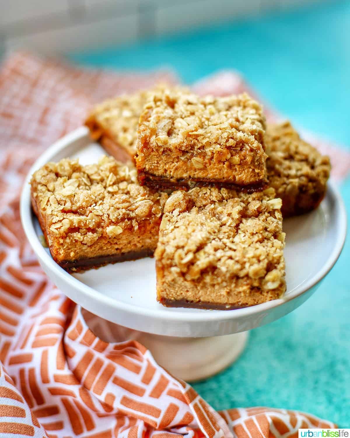 pumpkin cheesecake bars on a white cake stand with kitchen towel