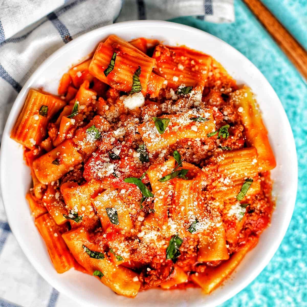 bowl of rigatoni pasta with Italian sausage on a white and blue checkered napkin and a fork.