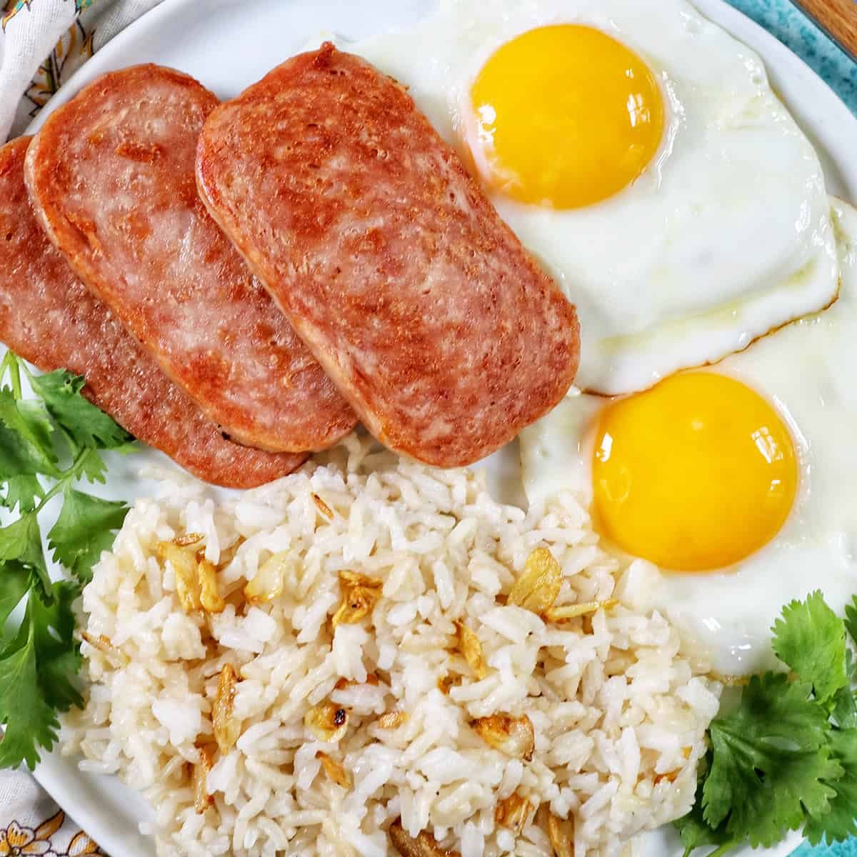 Plate of Spamsilog: slices of Spam, two eggs over simple, and Filipino Sinangag garlic rice with greens.