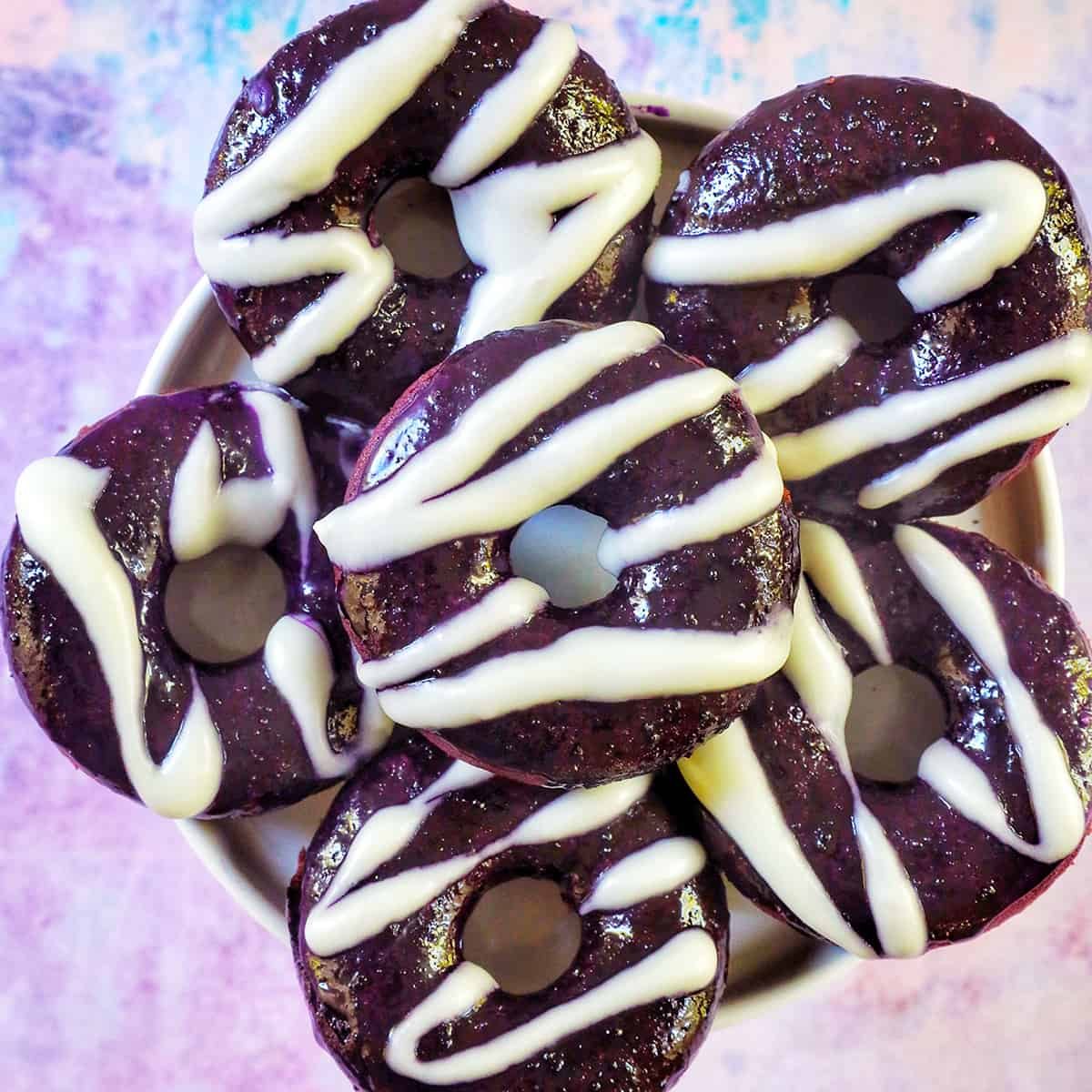 several ube donuts with ube glaze and white glaze drizzle on a purple and blue table.