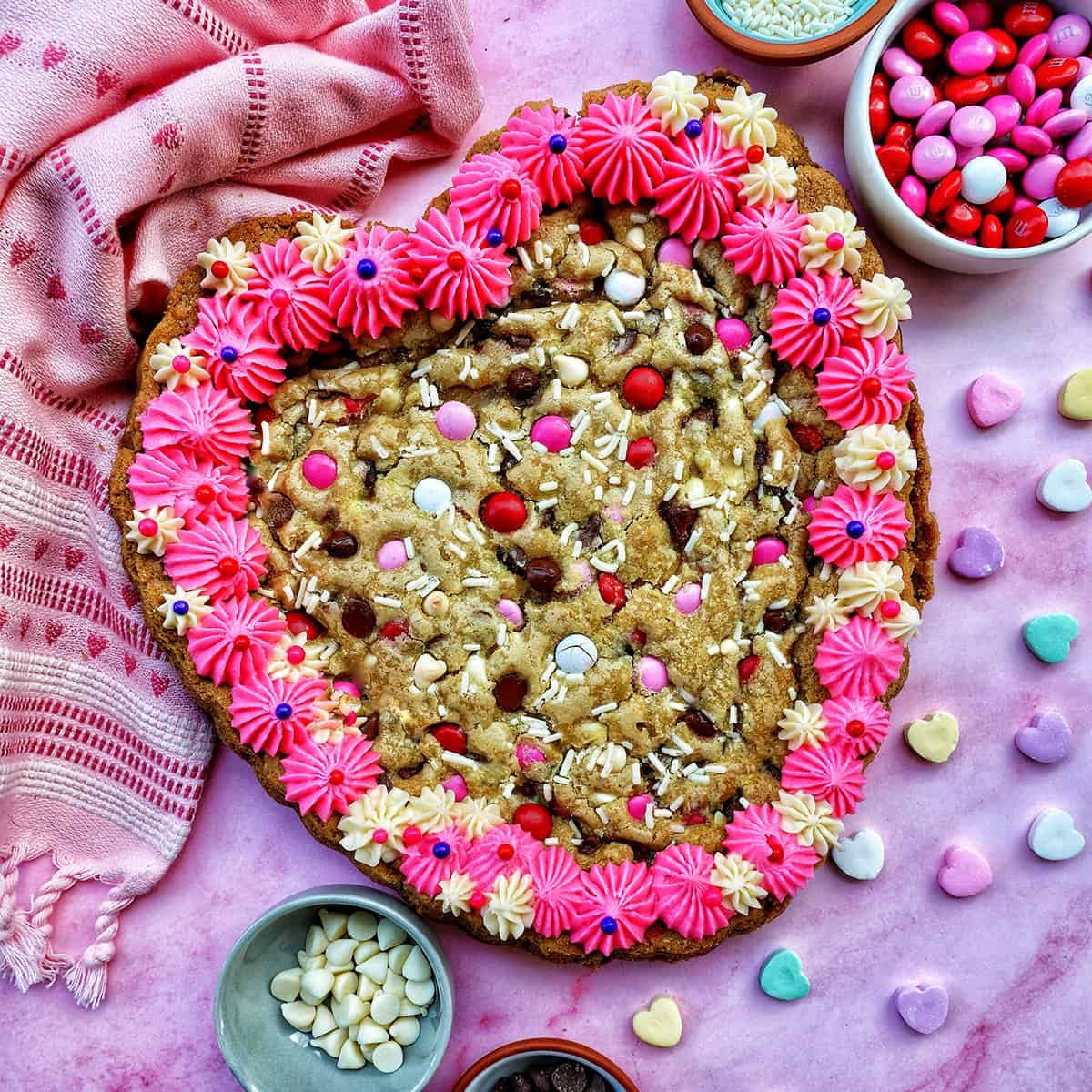 A Valentine's Day Cookie Cake with pink frosting, pink, red and white M & M candies, conversation hearts candies sprinkled around the