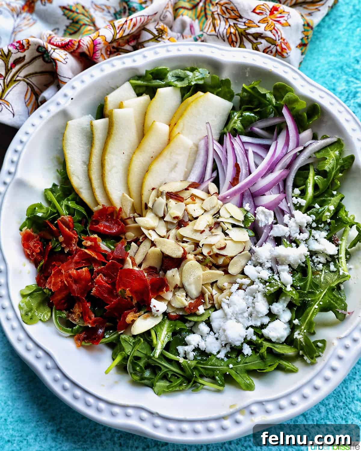 Sweet Pear and Peppery Arugula Salad 3 White bowl of arugula pear salad with red onions, dried cranberries, sliced almonds, and dairy-free feta cheese.