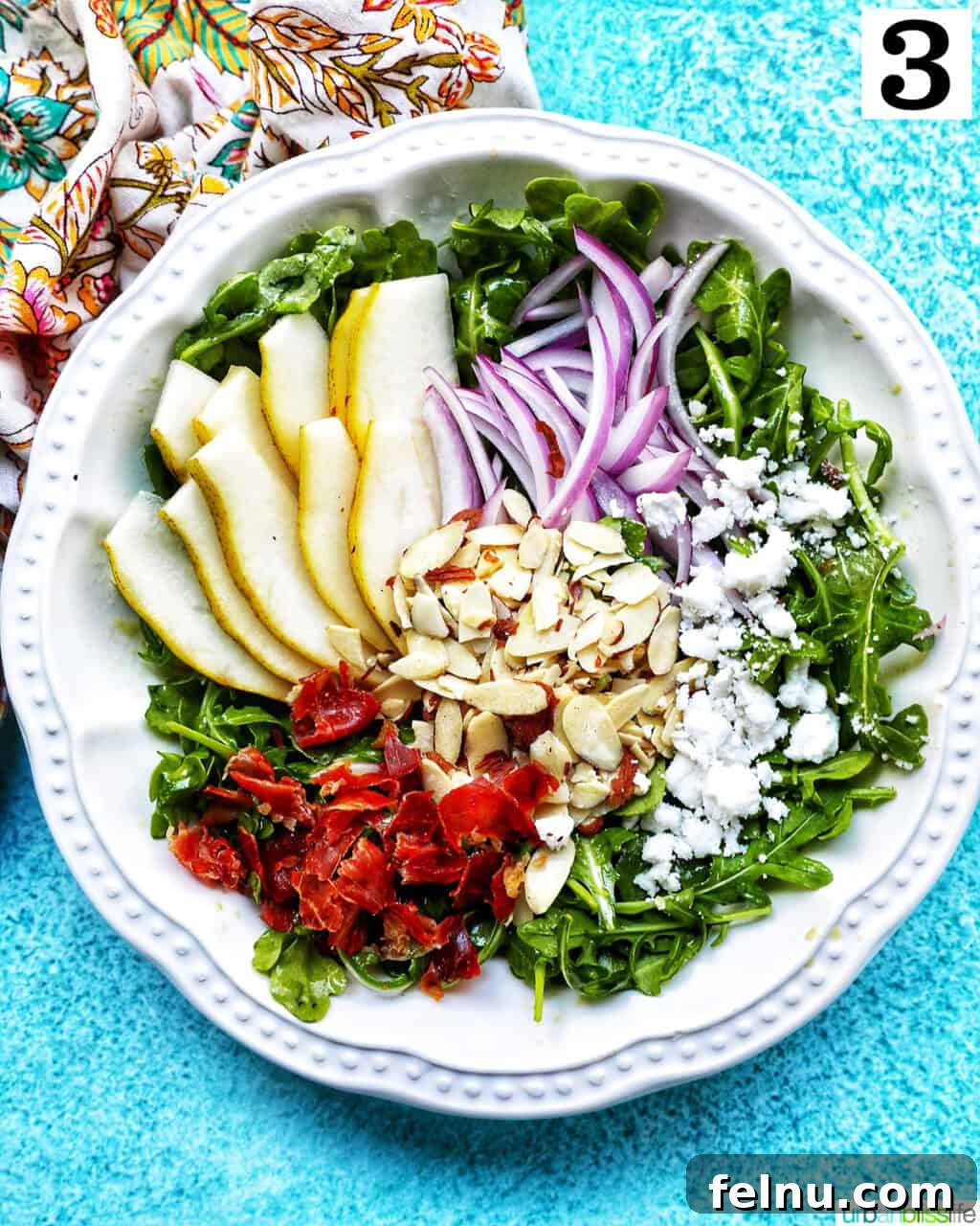 Sweet Pear and Peppery Arugula Salad 10 White bowl of finished arugula pear salad with red onions, dried cranberries, sliced almonds, and dairy-free feta cheese, tossed with dressing.