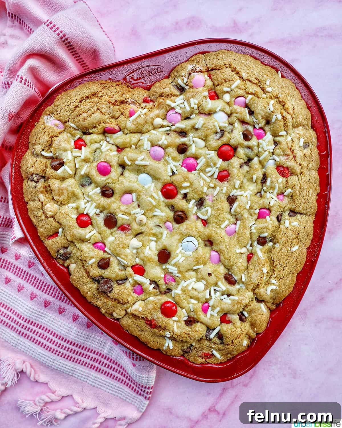 Love Struck Cookie Cake 3 a heart-shaped cookie cake baked in a red pan on pink background.