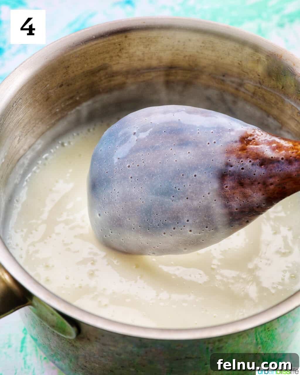 Thick white vanilla pudding clinging to the back of a wooden spoon, held above a saucepan of pudding.