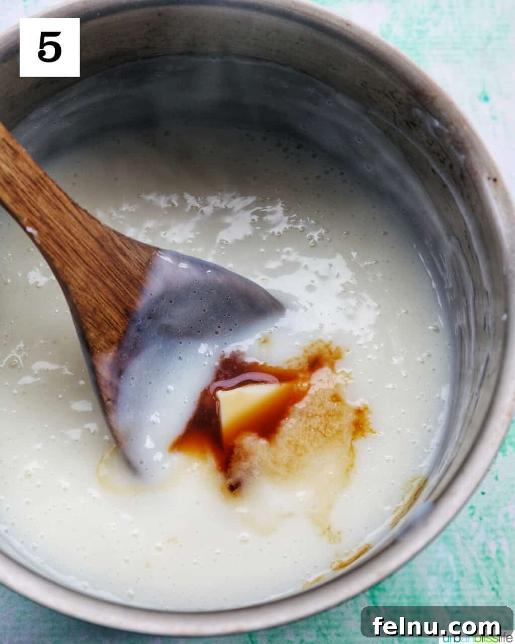 A wooden spoon stirring vanilla extract and butter into a saucepan of warm vanilla pudding.