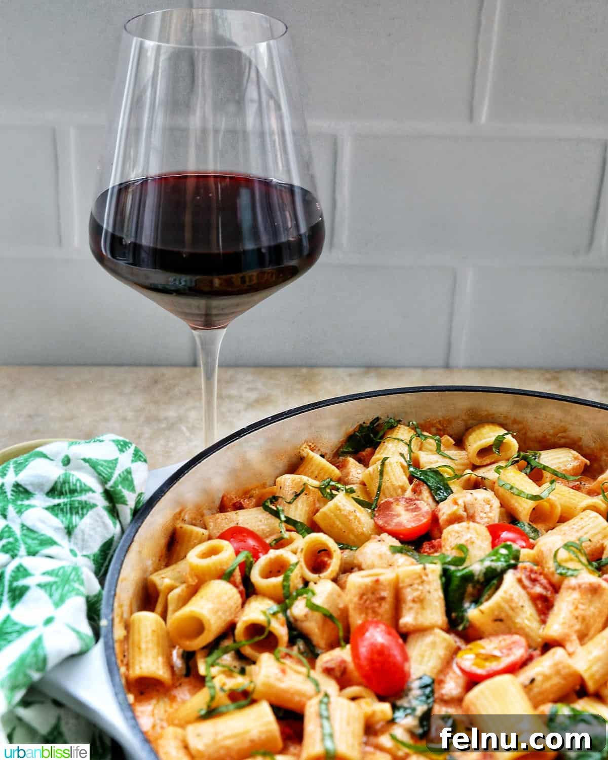 Velvety Tuscan Chicken Pasta 19 A glass of red wine next to a large skillet of creamy Tuscan chicken pasta, creating an inviting meal scene.