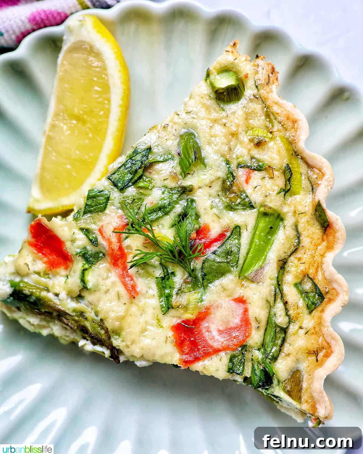 Smoked Salmon & Dill Quiche 3 A delectable slice of Salmon Quiche on a plate, featuring a golden crust, tender asparagus, and leeks, accompanied by a fresh lemon slice.