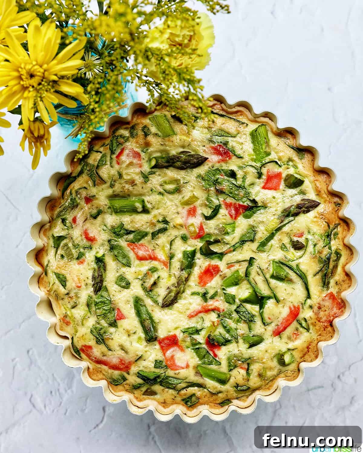 Smoked Salmon & Dill Quiche 21 A beautifully baked Salmon Quiche in a specialized pie pan, adorned with salmon, asparagus, and leeks, presented in front of a vase of cheerful yellow flowers.