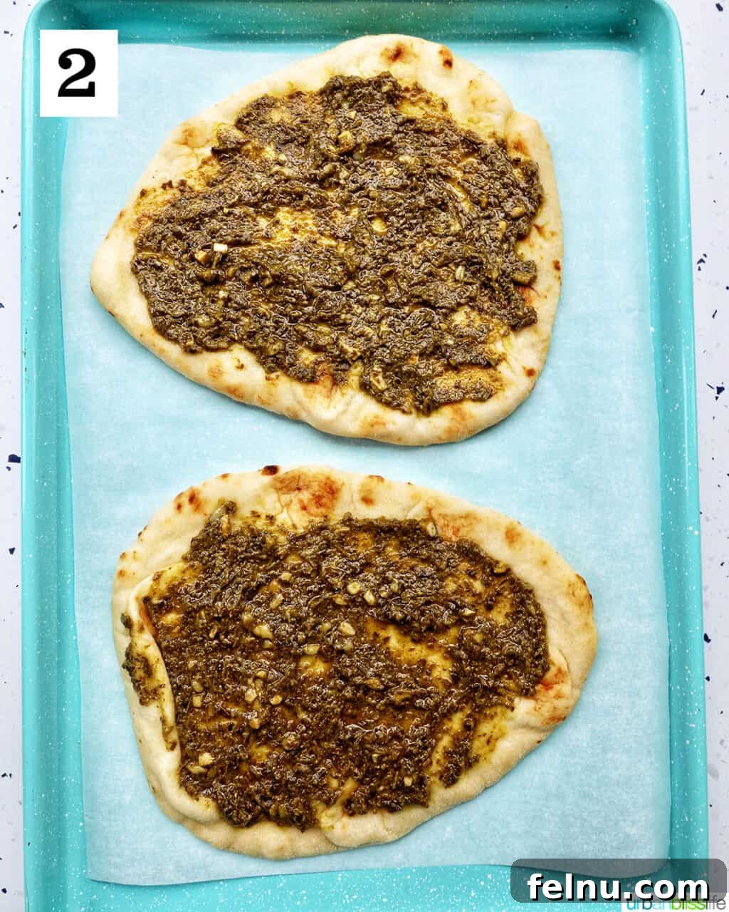 two naan flatbreads on a baking sheet, generously spread with vibrant green pesto sauce.