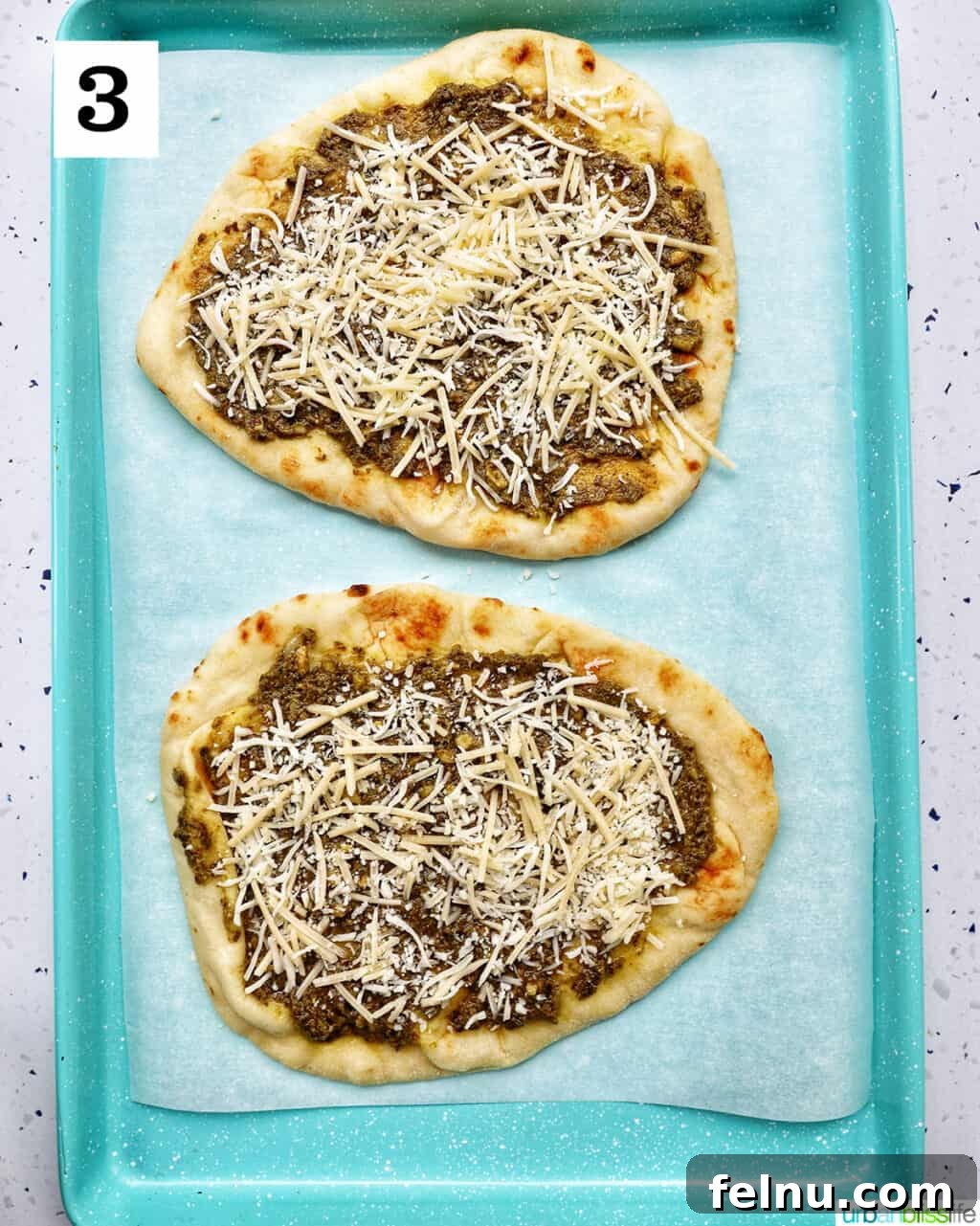 two naan flatbreads on a baking sheet, layered with pesto and sprinkled with shredded mozzarella cheese.