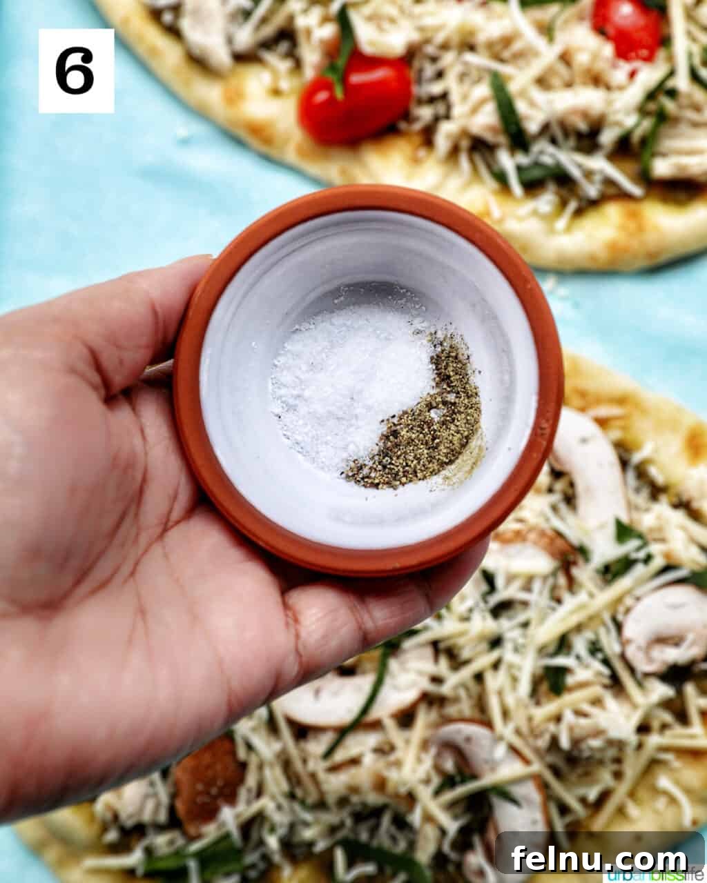 A hand gently sprinkling salt and pepper over a prepared chicken pesto flatbread, adding the finishing seasonings.