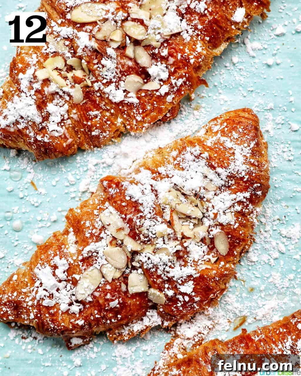 Three almond croissants on a baking sheet, fully topped with powdered sugar and sliced almonds.