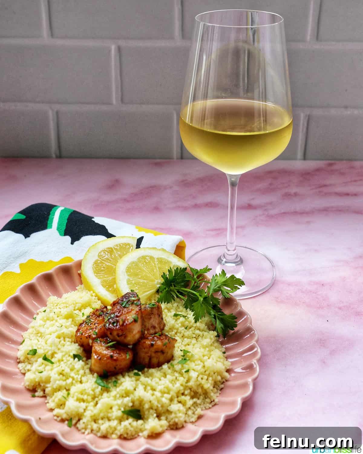 Cast iron seared scallops on a bed of couscous garnished with herbs and lemon slices and a glass of white wine.