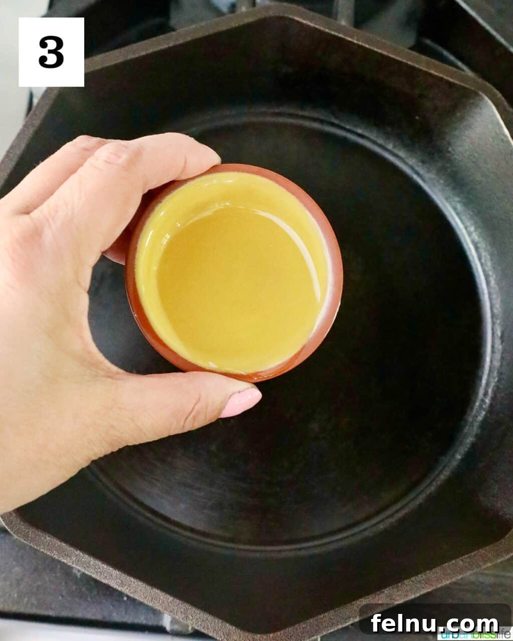 Hand holding a small bowl of olive oil over a cast iron skillet.