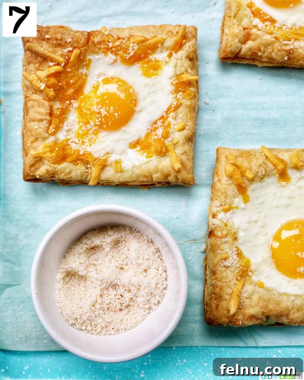 Golden Egg Parcels 13 Baked eggs in puff pastry on a baking sheet, with a small bowl of grated Parmesan cheese nearby for garnish.