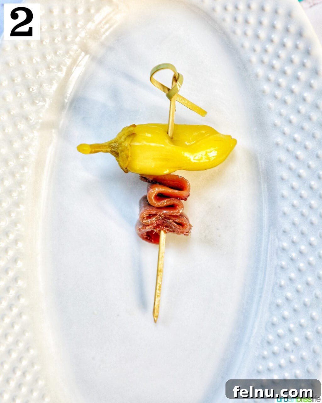 Gilda A Basque Classic 6 White plate with a single food pick featuring a pepper and a folded anchovy, showing the second step of Gilda pintxo assembly.