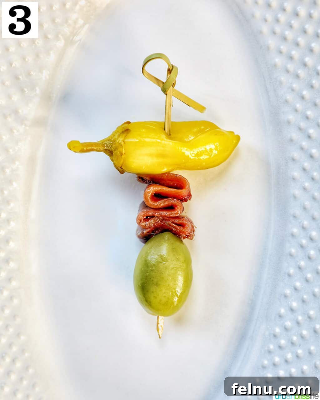 Gilda A Basque Classic 7 White plate with a completed Gilda pintxo on a food pick, featuring a pepper, an anchovy, and an olive.