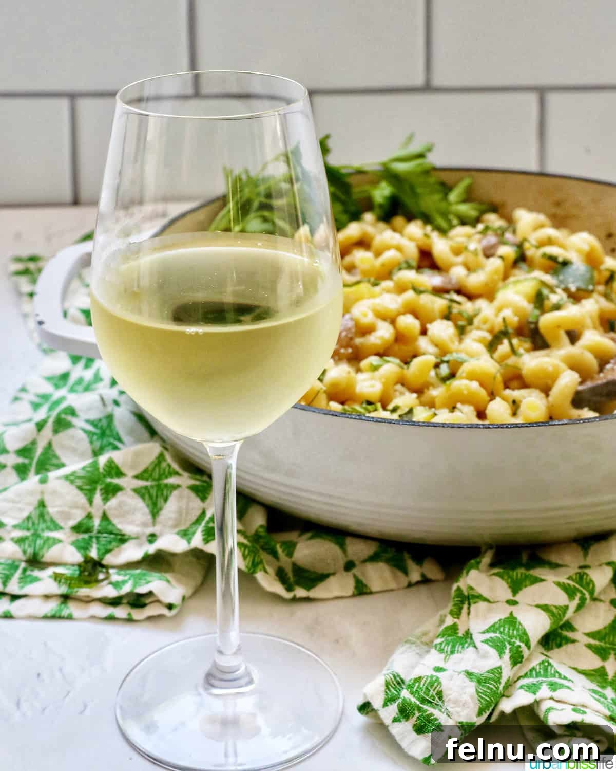 An elegant glass of white wine poised in front of a meticulously prepared pan of pasta, accompanied by a tasteful green and white napkin.