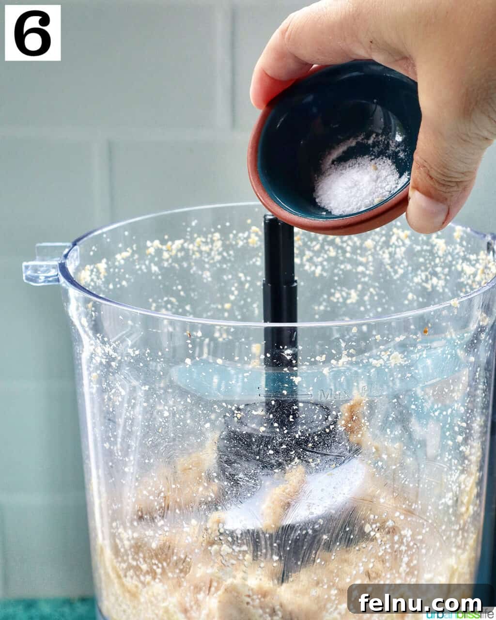 Crafting Your Own Tahini 12 hand holding salt over a food processor with sesame seeds making tahini.