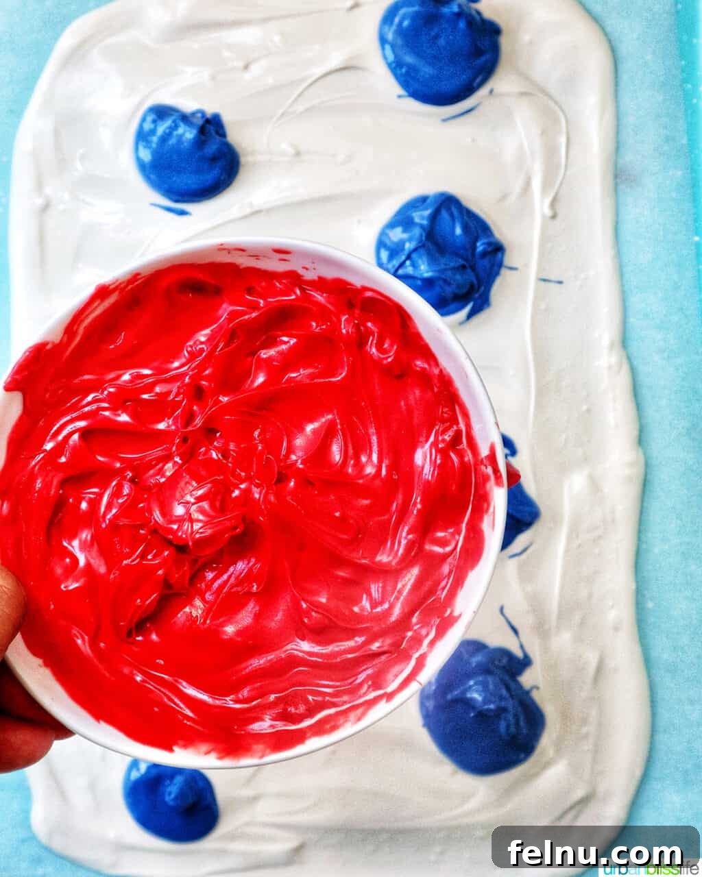Fourth of July Freedom Bark 7 A hand carefully holding a bowl of rich red melted chocolate, ready to drizzle it over the white and blue base on the baking sheet.