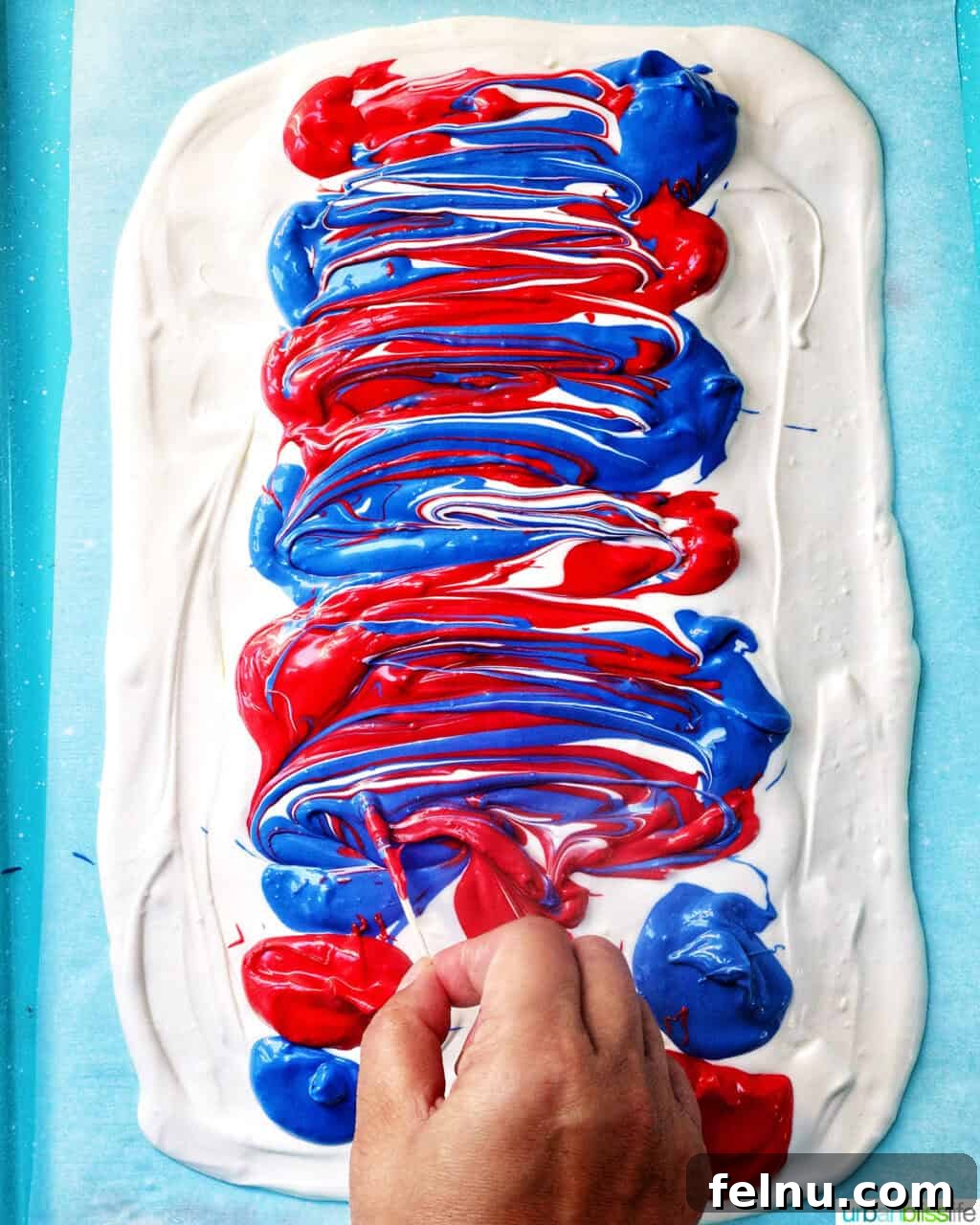 Fourth of July Freedom Bark 9 A hand holding a toothpick, delicately creating intricate designs and swirls in the melted red, white, and blue chocolate mixture on the baking sheet.