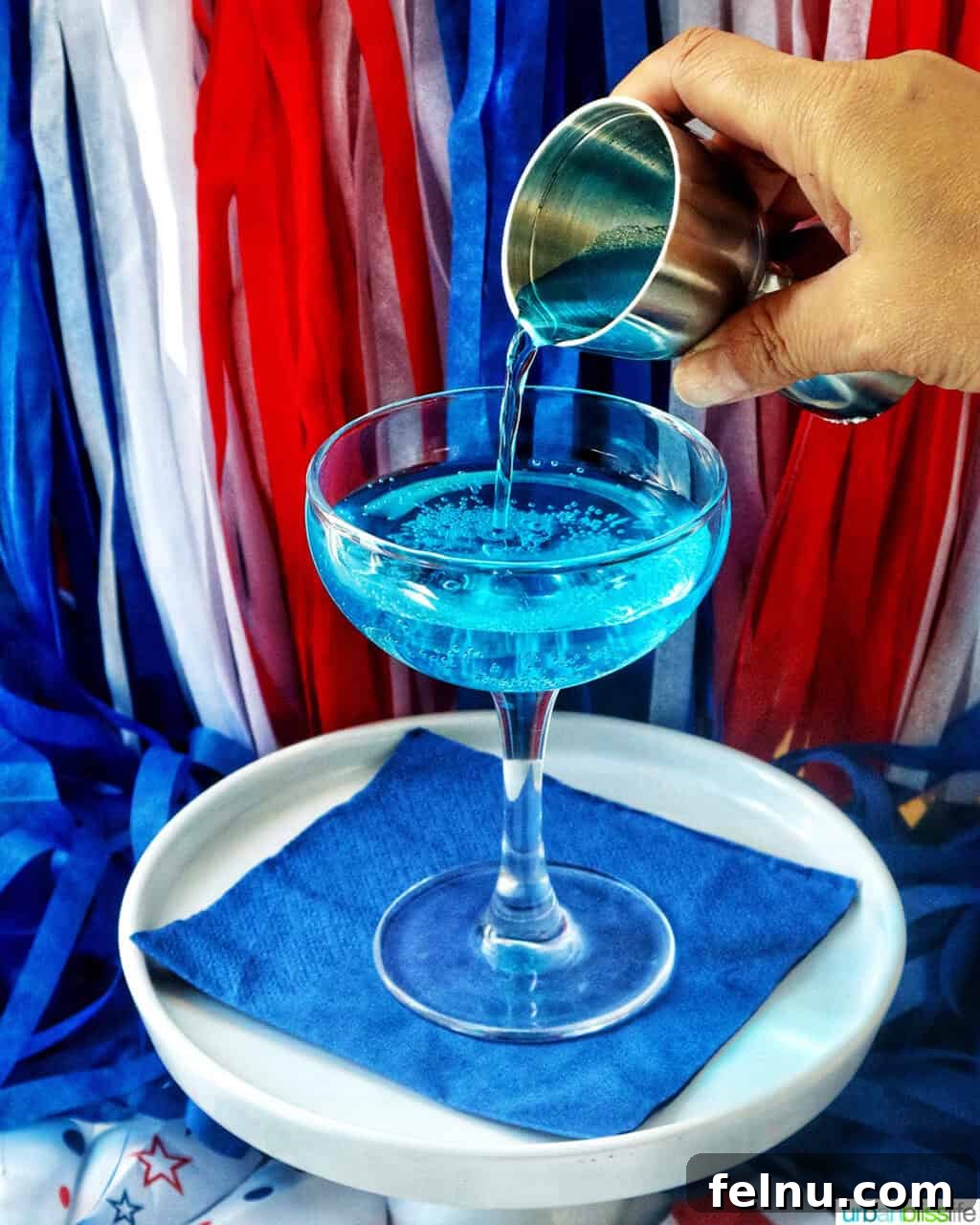 Pouring curacao into a cocktail glass with red, white, and blue decorations in the background.