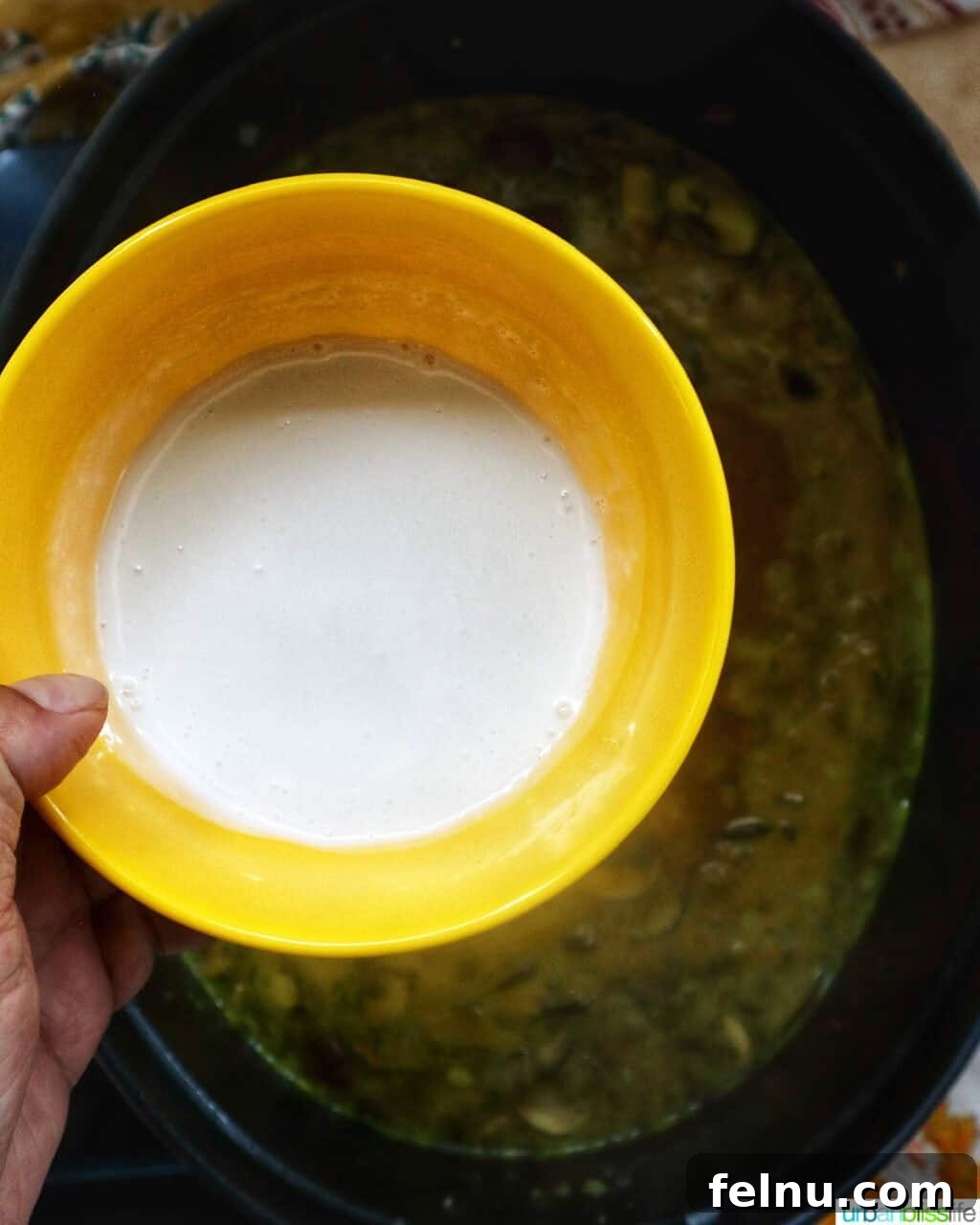 Smoky Sausage Pumpkin Bisque 11 A hand carefully pouring rich whipping cream from a yellow bowl into a simmering Dutch oven of pumpkin soup, adding luxurious creaminess.