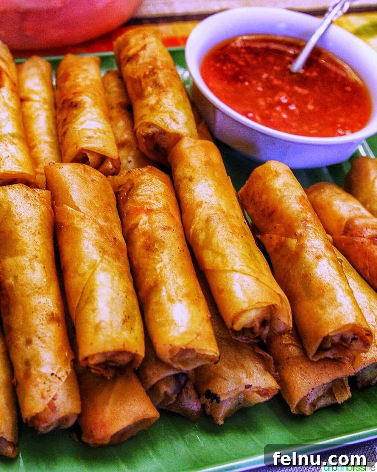 Tray of golden brown Filipino lumpia egg rolls, stacked alongside a small bowl of sweet chili dipping sauce.