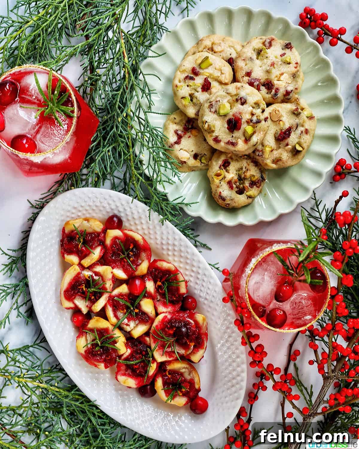 Cranberry brie tartlet appetizers on a plate next to a plate of pistachio cranberry shortbread cookies and glasses of cranberry mocktails with holiday decor
