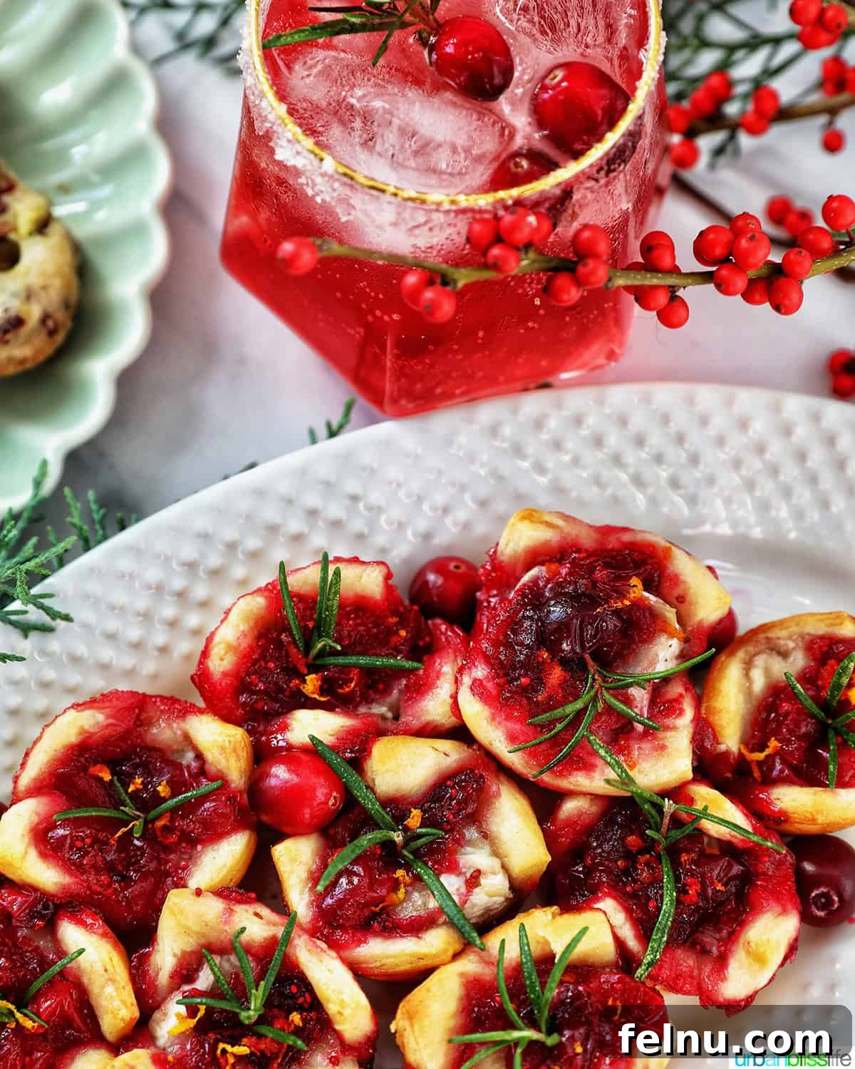 Cranberry brie tartlet appetizers on a plate next to a plate of pistachio cranberry shortbread cookies and glasses of cranberry mocktails with holiday decor