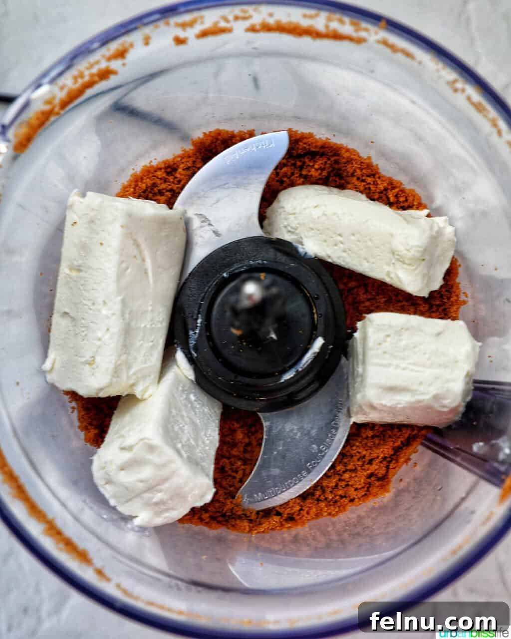 Biscoff Delights 6 Cream cheese being added to the Biscoff cookie crumbs inside a food processor.