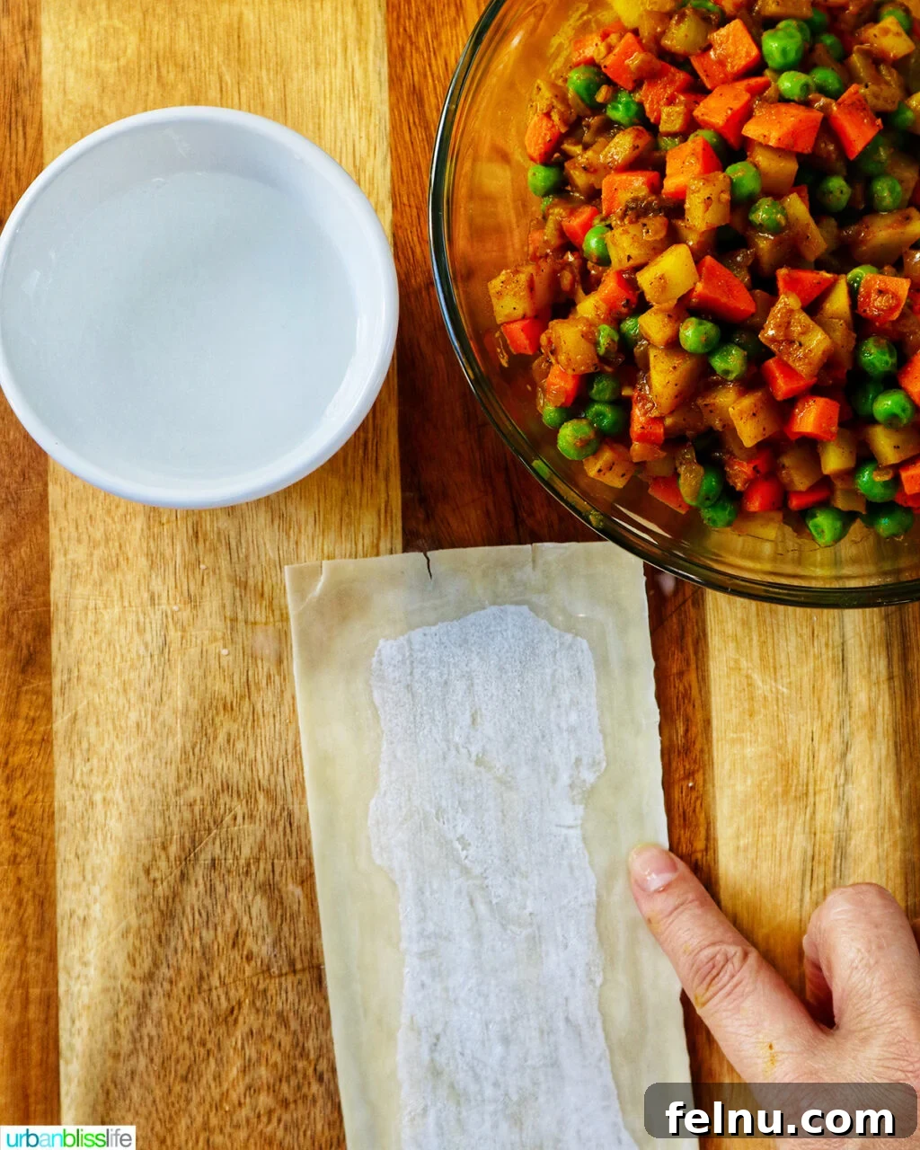 Hand wetting the sides of an egg roll wrapper on a cutting board with bowl of curry samosa filling.
