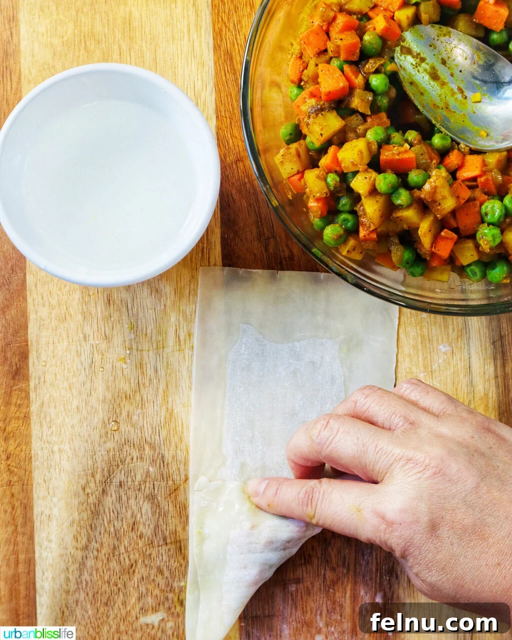 Hand folding curry samosa wrapper on a cutting board with bowl of curry samosa filling.