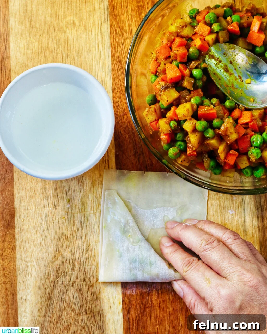 Hand folding curry samosa wrapper on a cutting board with bowl of curry samosa filling.