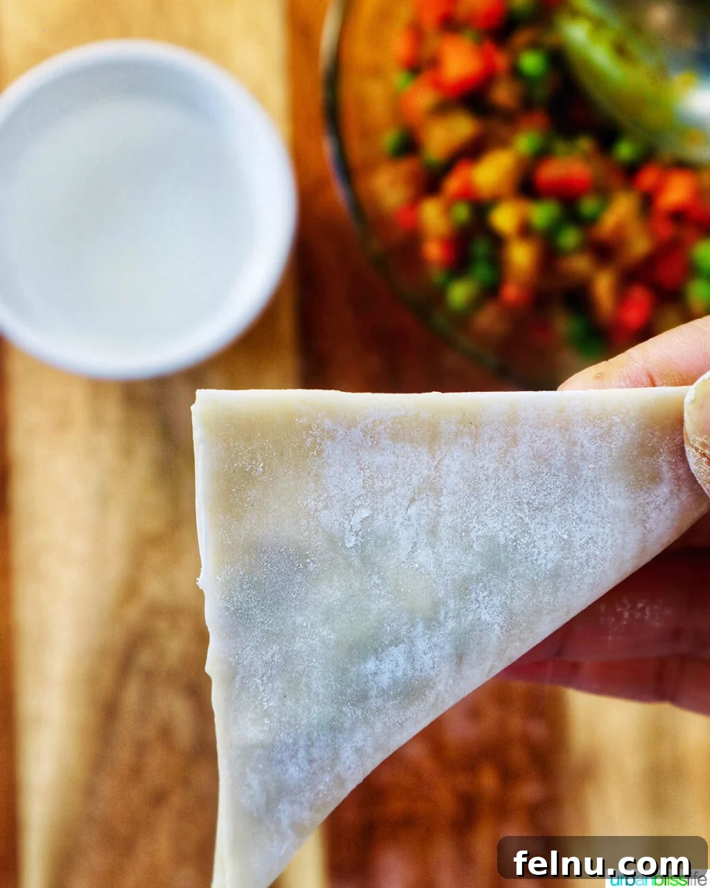 Hand folding curry samosa wrapper on a cutting board with bowl of curry samosa filling.