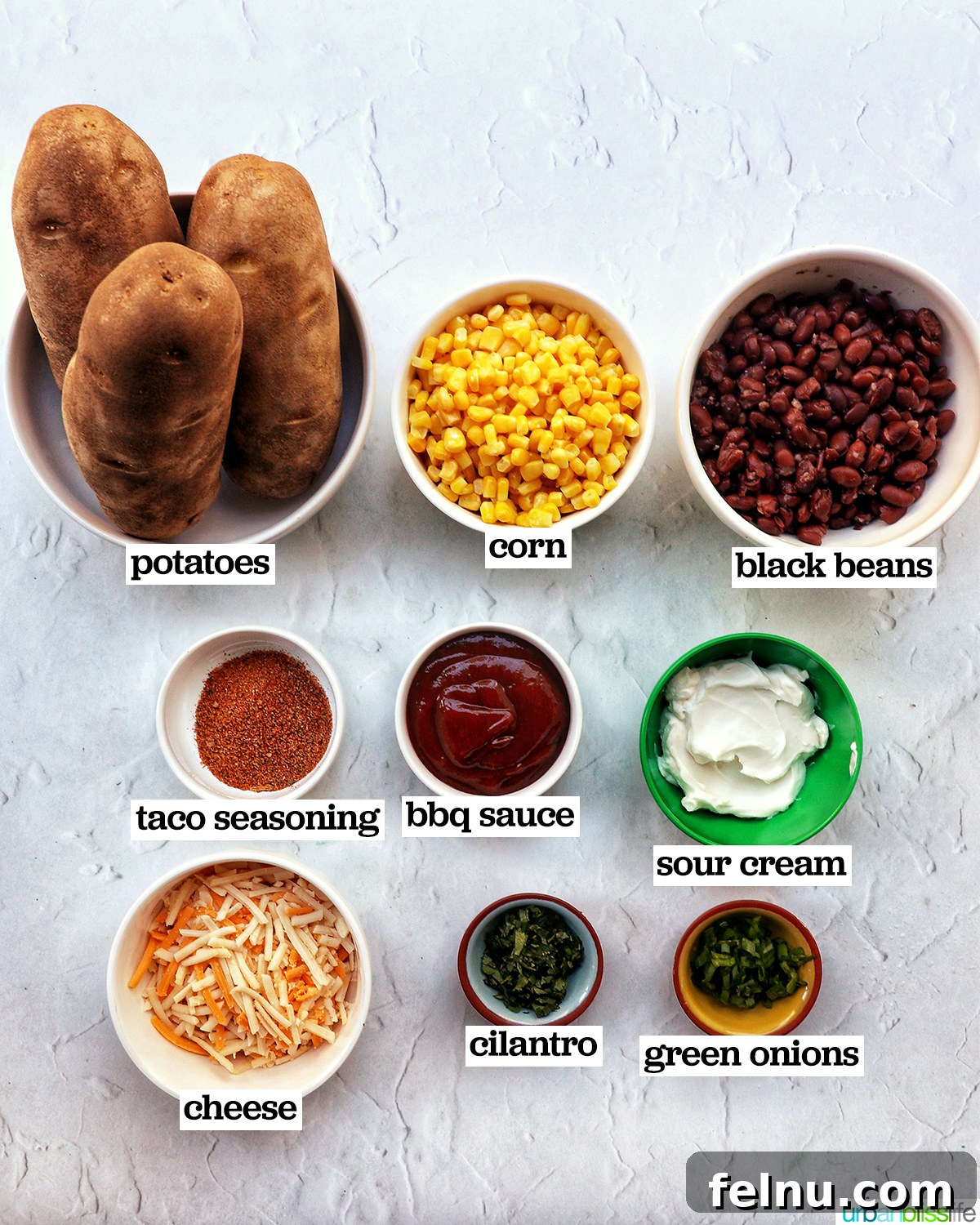 Hearty Spud Indulgence 4 A collection of ingredients for loaded baked potato bowls, including russet potatoes, a bowl of frozen corn, a can of black beans, olive oil, taco seasoning, shredded cheese, barbecue sauce, sour cream, and sliced green onions.