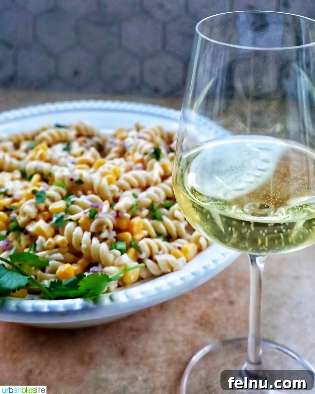 Zesty Elote Pasta Salad 18 Mexican street corn pasta salad in a pristine white bowl, elegantly accompanied by a tall glass of chilled white wine, set against a warm, inviting backdrop.