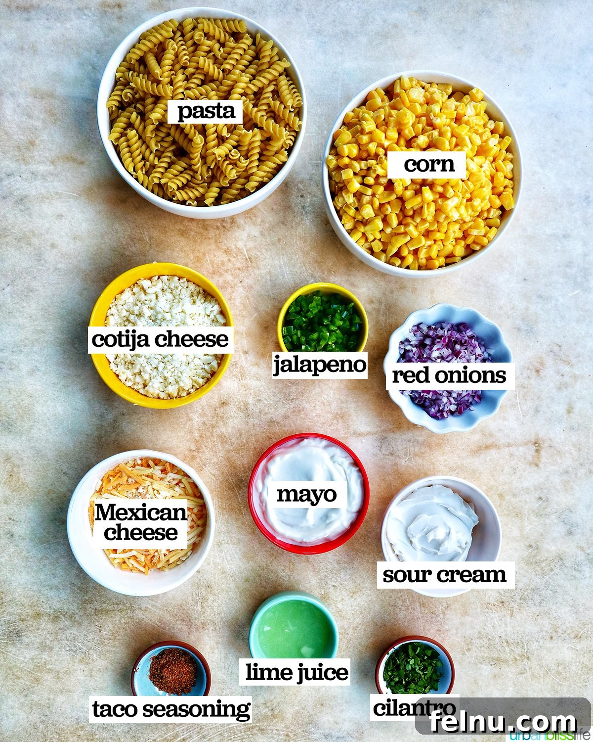 Zesty Elote Pasta Salad 4 A flat lay of fresh, colorful ingredients meticulously laid out on a kitchen surface for Mexican street corn pasta salad, including pasta, corn, a variety of cheeses, red onion, jalapeño, and aromatic herbs.