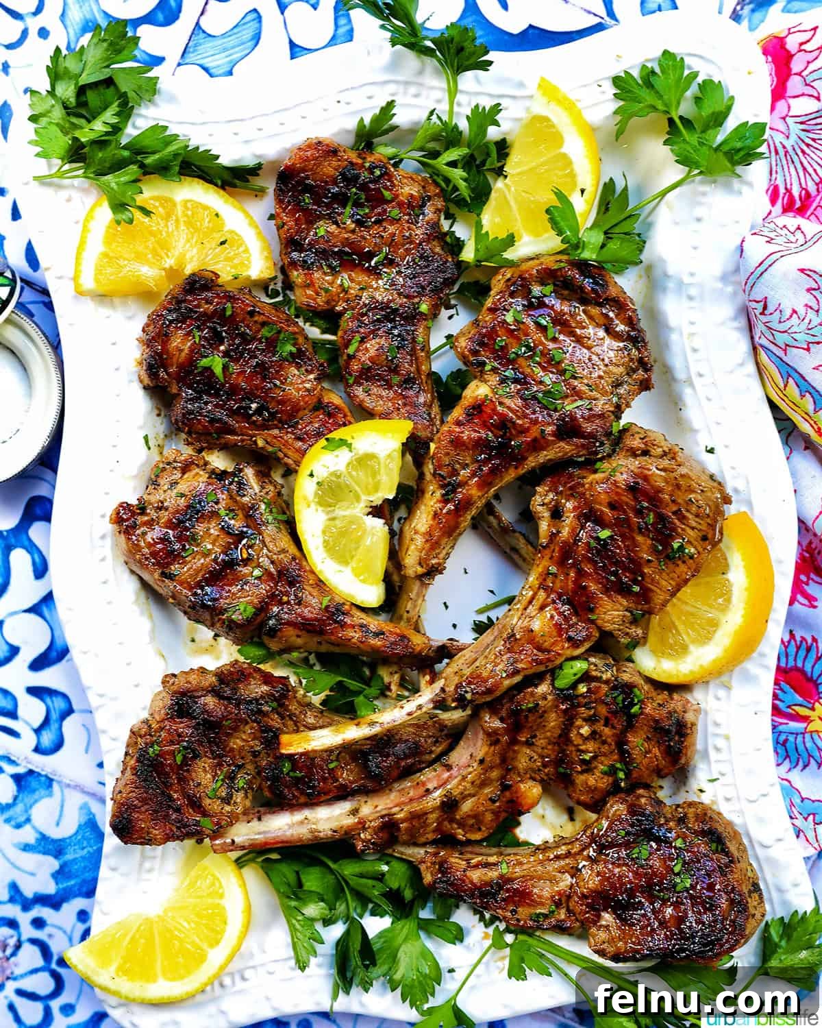 Aegean Lamb Chops 8 Pile of Greek lamb chops on a white plate with fresh herbs and lemon wedges.