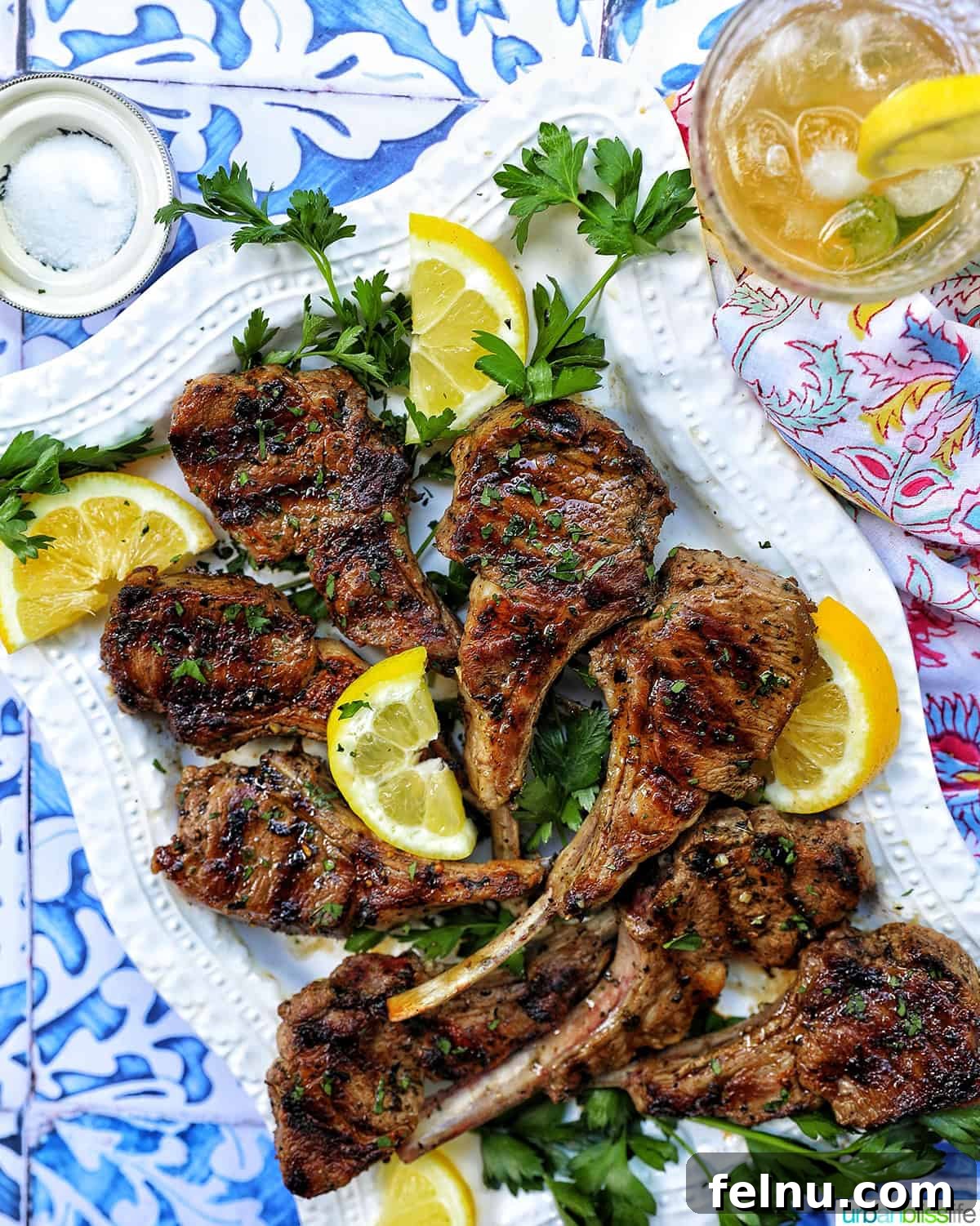Aegean Lamb Chops 10 Greek lamb chops on a white plate with herbs, lemon slices, and a glass of Bourbon Lemonade cocktail.
