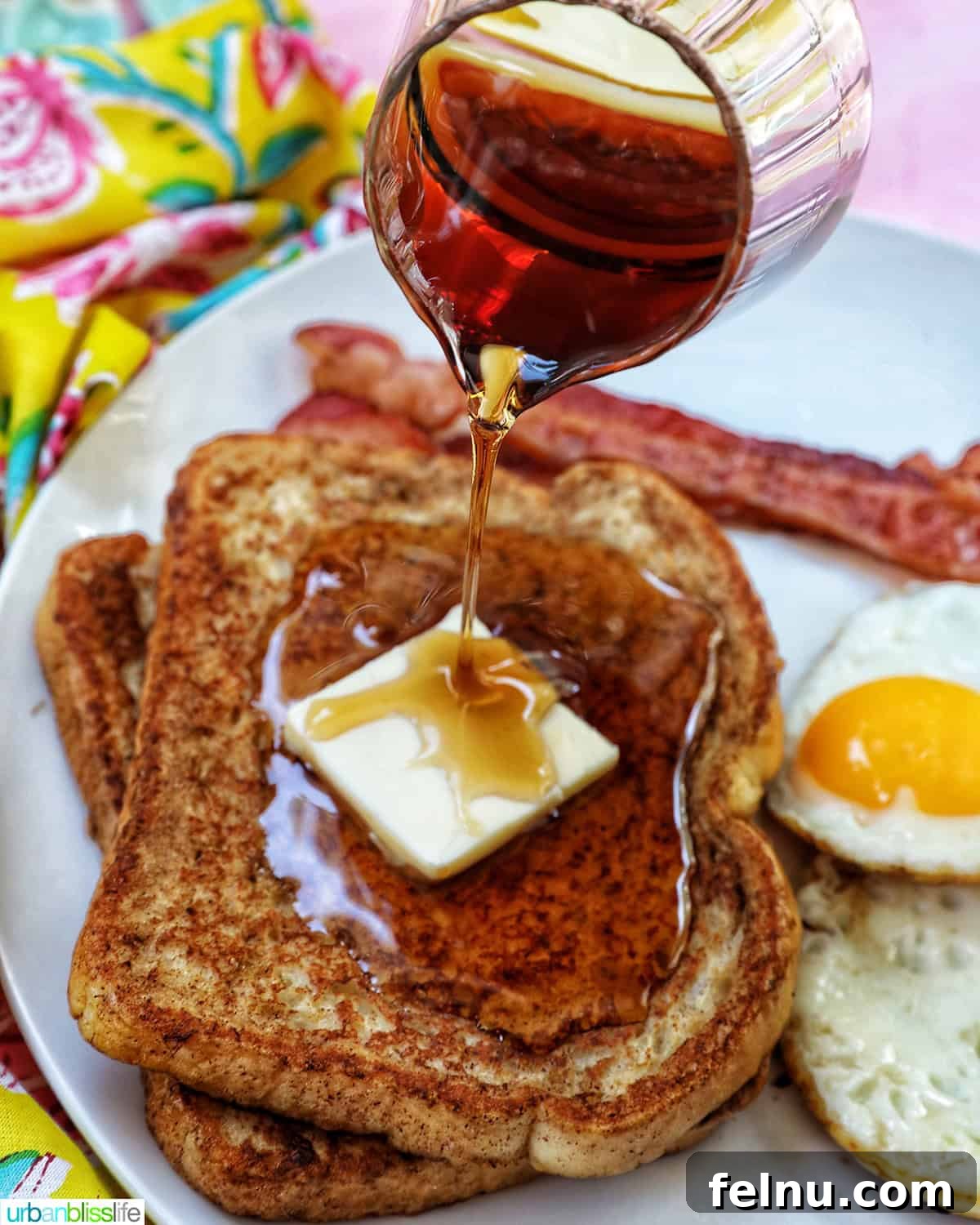 Power-Packed Protein French Toast 3 maple syrup pouring over a stack of high protein french toast with bacon and eggs on the side.